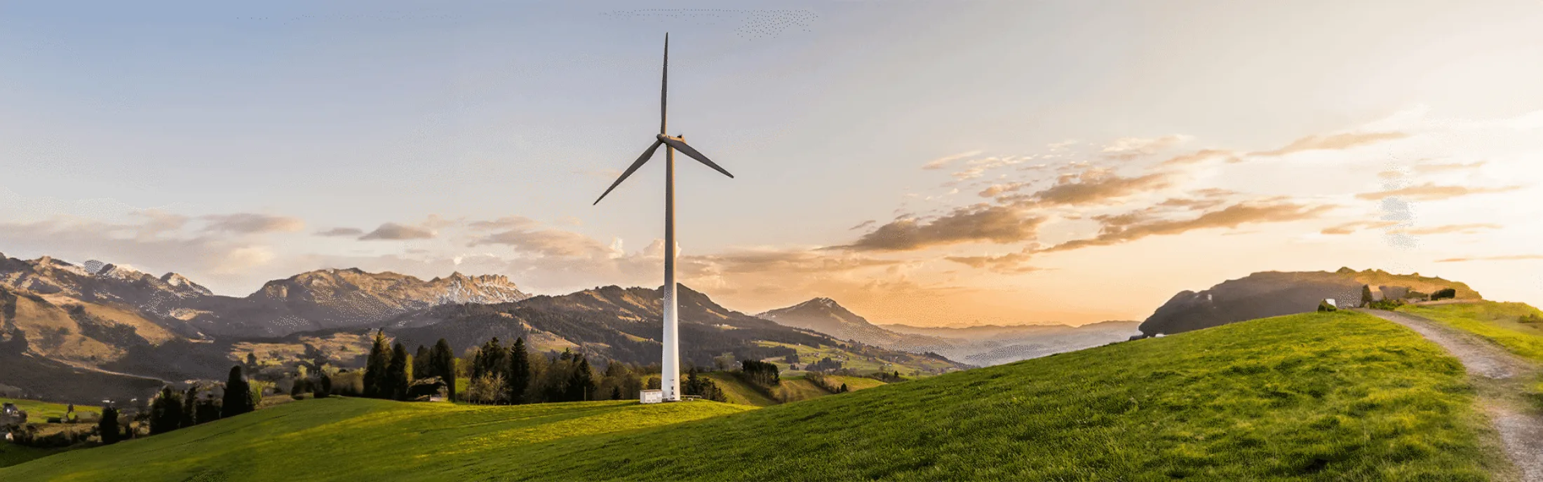 Wind turbine on a green hillside with mountains in the background, set against a sunset sky.