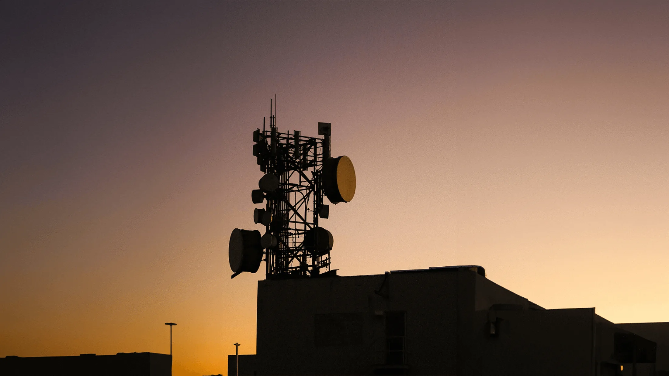 Silhouette of a cell tower with multiple antennas and dishes on top of a building, set against a warm sunset sky.