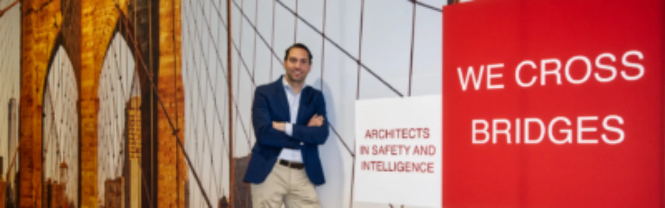 A man in a blue blazer standing in front of a backdrop featuring a bridge and the words 'We Cross Bridges' and 'Architects in Safety and Intelligence' displayed in bold red and white colors.