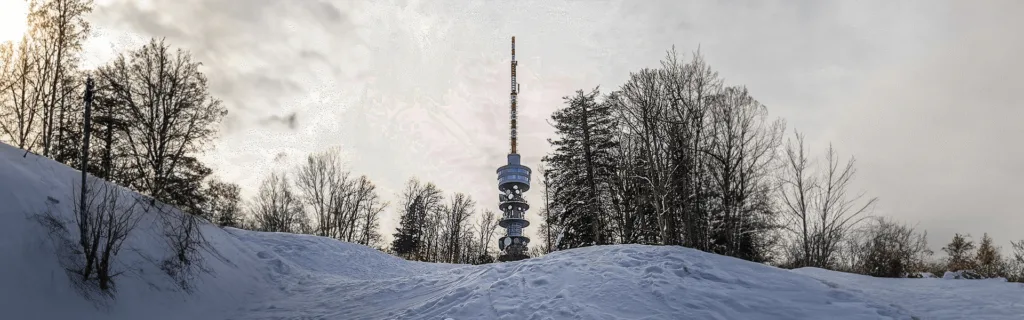 Telecommunication tower surrounded by a snowy landscape and bare trees under a cloudy sky.