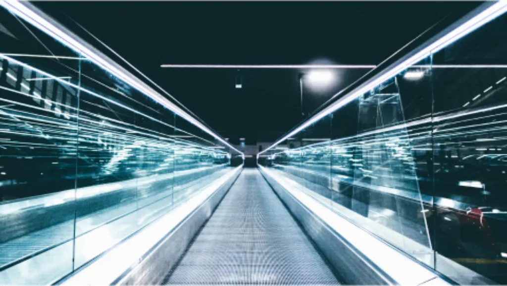 A long, illuminated walkway with reflective glass walls creating a tunnel-like effect, giving an impression of high speed and motion.