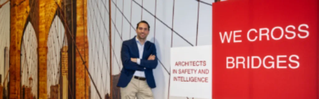 A man in a blue blazer standing in front of a backdrop featuring a bridge and the words 'We Cross Bridges' and 'Architects in Safety and Intelligence' displayed in bold red and white colors.